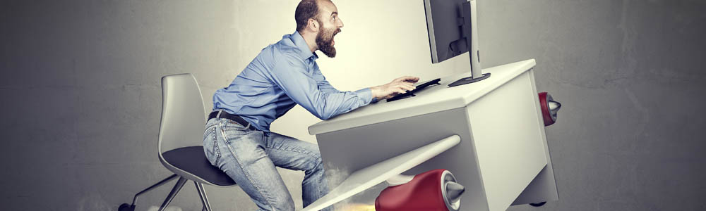 Man sitting at his desk, which has rockets on its legs and is about to take off. Man with a beard in a blue dress shirt is screaming while looking at his computer screen.